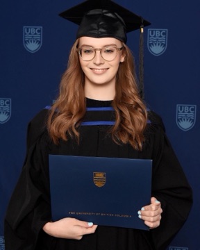 Elisha in a black graduation cap and gown and round glasses, smiling and holding a navy University of British Columbia diploma folder against a UBC-branded backdrop.